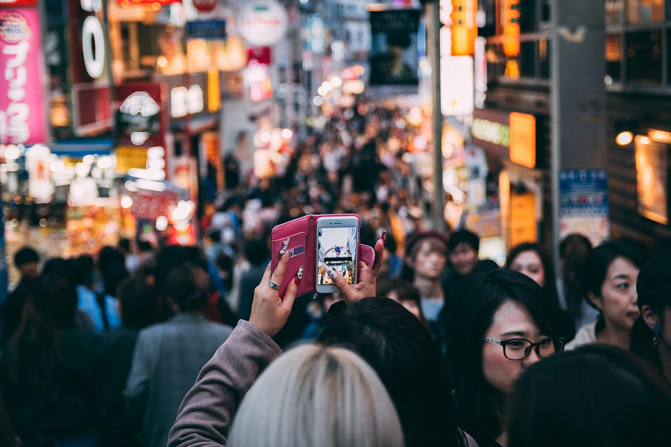 A woman in a crowded urban street, raises her hands holding an iPhone to take a photo.