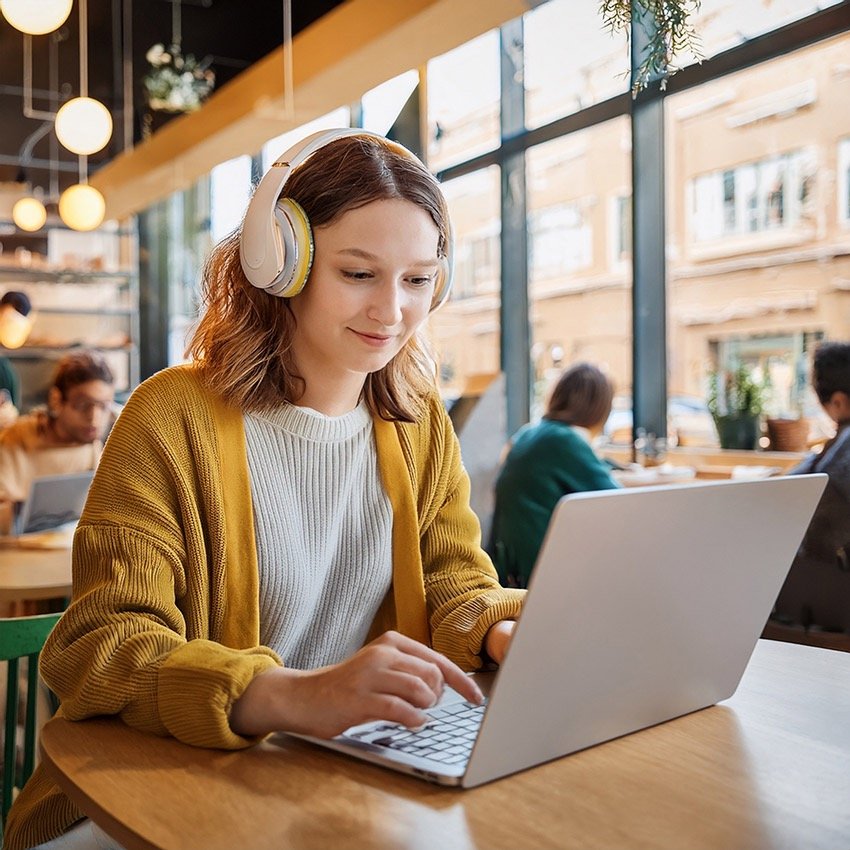 A young woman is sitting in a coffee shop, focussing and typing on an open laptop in front of her, wearing headphones.