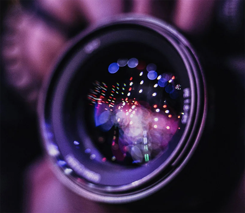 Close-up of a camera lens held to take a shot, with the street lights reflecting in it.