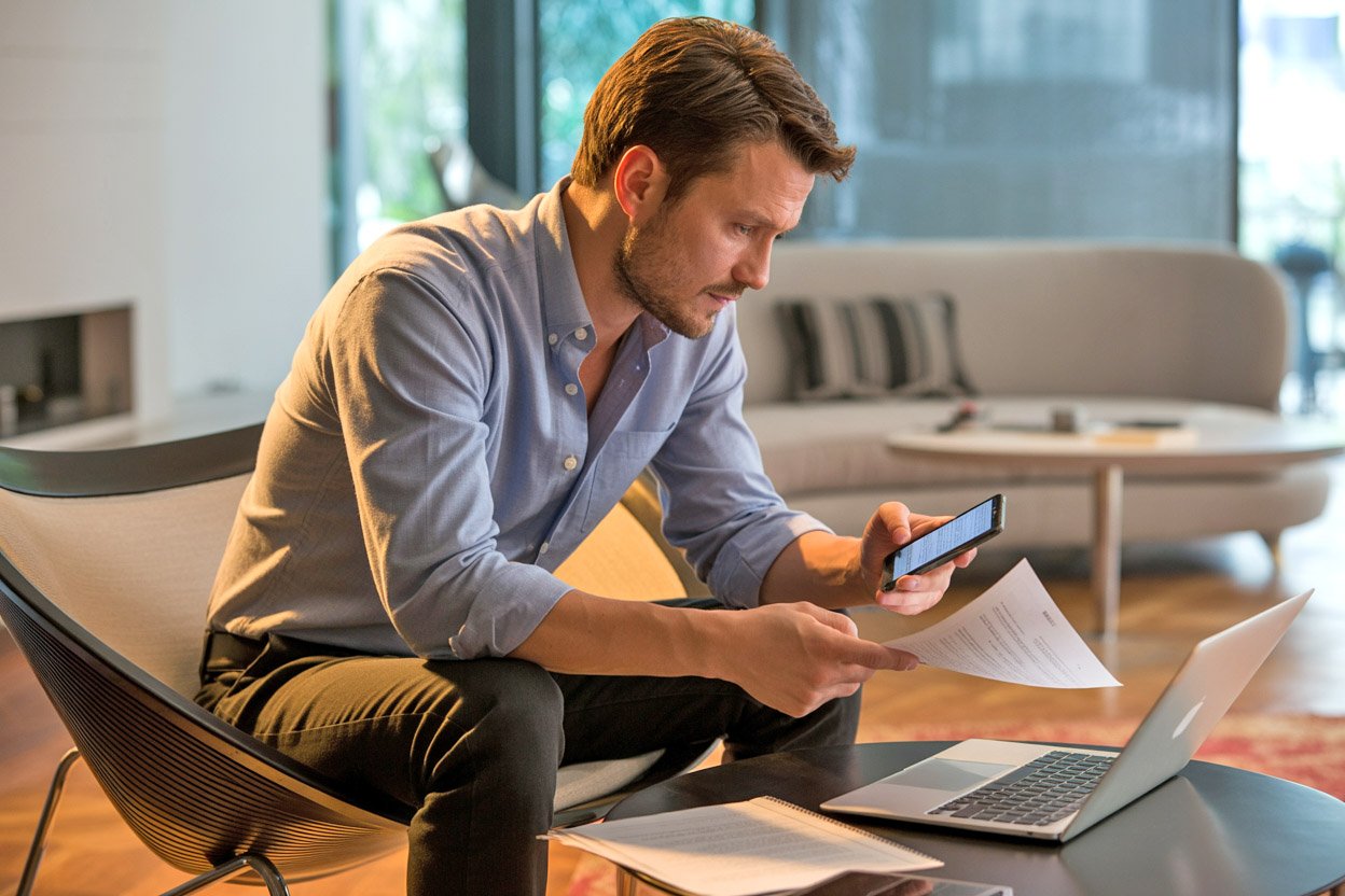A man sitting in front of a laptop at home, checking facts on his phone and with documents in his hands.